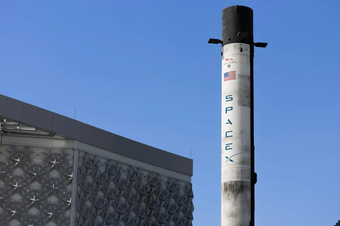 SpaceX rocket displayed vertically near a modern building with a textured facade, set against a clear blue sky, conveying innovation and technology.