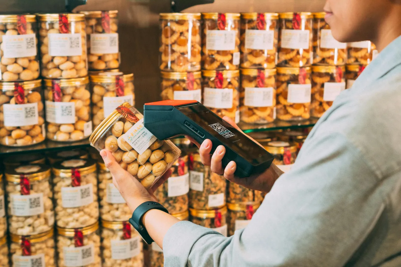 A person scans a jar of cookies with a handheld device in a store. Shelves are filled with identical jars, creating an organized and commercial atmosphere.