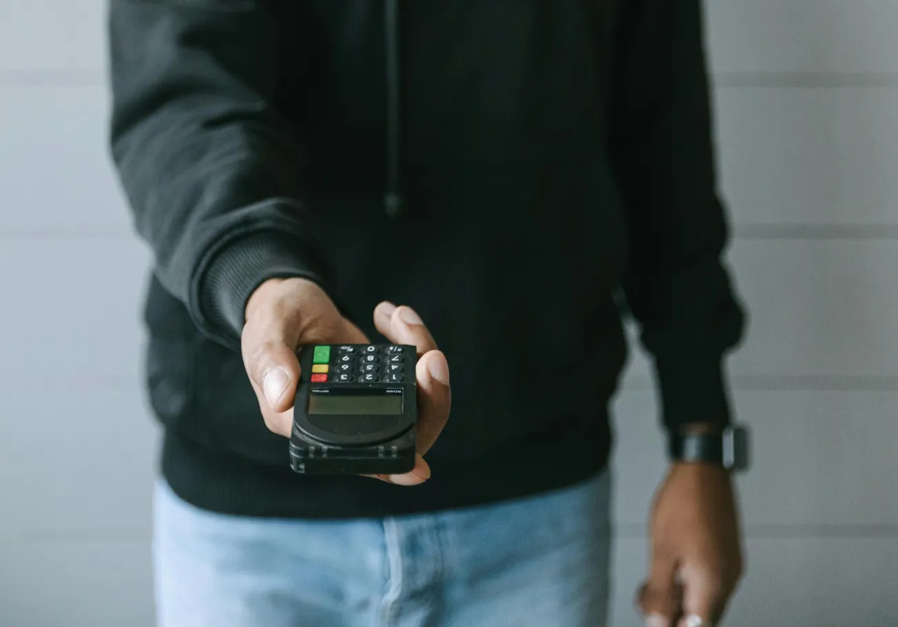 A person in casual attire holds out a payment terminal, inviting a transaction. The device's colorful buttons stand out, conveying a sense of modern commerce.
