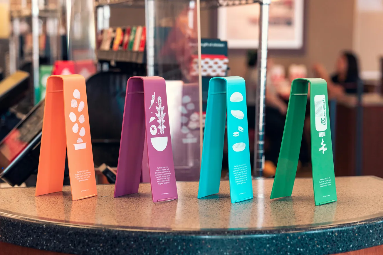 Four colorful, tent-shaped signs on a counter display food illustrations and text. The vibrant orange, purple, teal, and green signs add a lively touch.