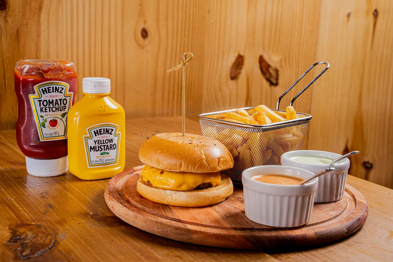 Burger with melted cheese on a wooden plate, accompanied by fries in a metal basket, two sauces, and bottles of ketchup and mustard on a wooden table.