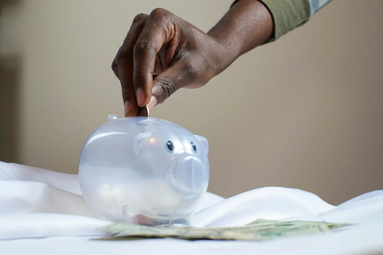 A hand drops a coin into a clear piggy bank, symbolizing saving money. Bills are scattered below on a white surface, conveying financial planning.
