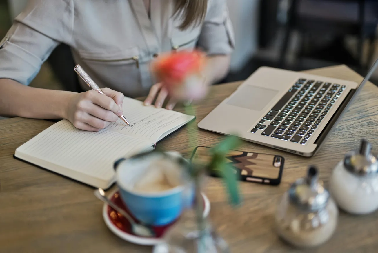 A person writes in a notebook beside a laptop on a wooden table with a blurred coffee cup and flower in the foreground, creating a cozy, focused atmosphere.
