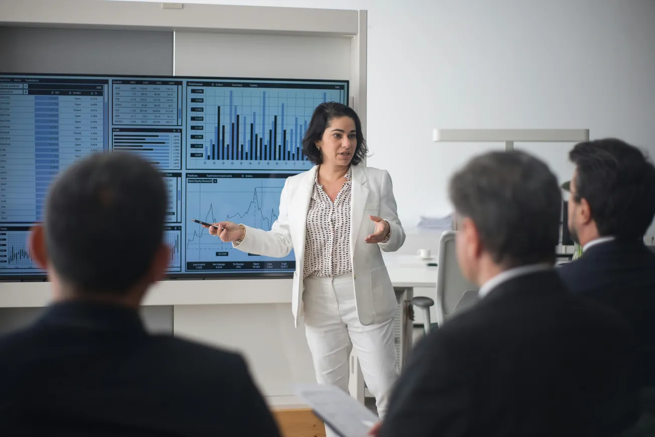 A woman in a white suit presents data on wall screens to an audience of three men in suits. Charts and graphs indicate a professional setting.