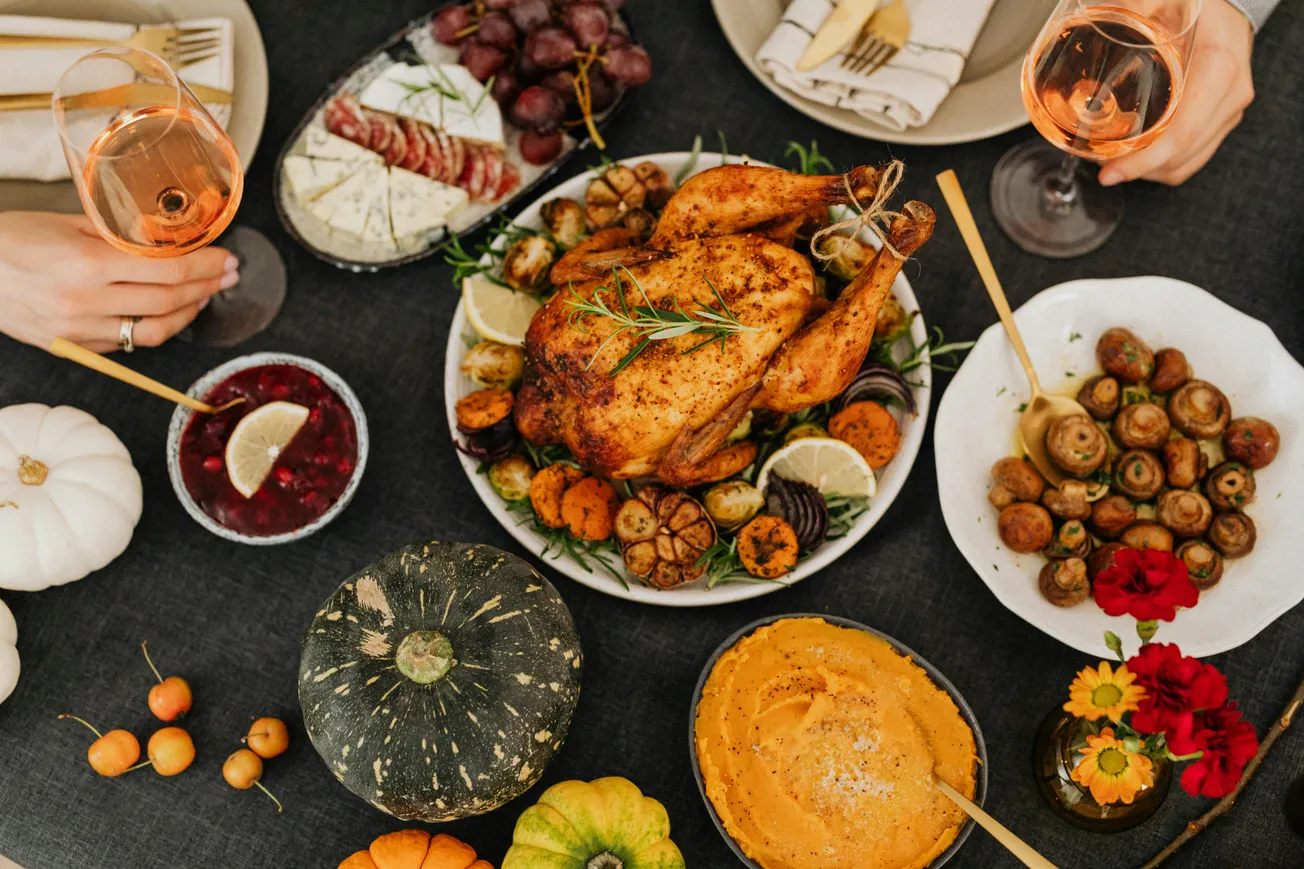 A Thanksgiving feast features a roasted turkey surrounded by vegetables, cheese platter, mushrooms, cranberries, and glasses of rosé wine, evoking warmth.