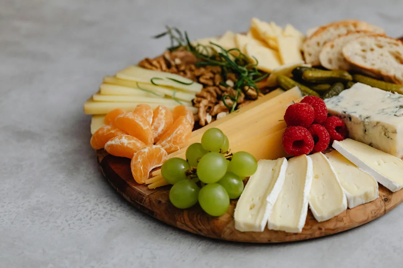A round wooden board displays assorted cheeses, grapes, raspberries, orange slices, nuts, pickles, and baguette slices. The scene is appetizing and elegant.
