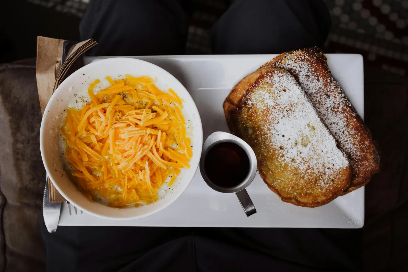 A rectangular plate on a lap holds a bowl of shredded cheese, powdered French toast, syrup, and napkin. The setting feels cozy and inviting.