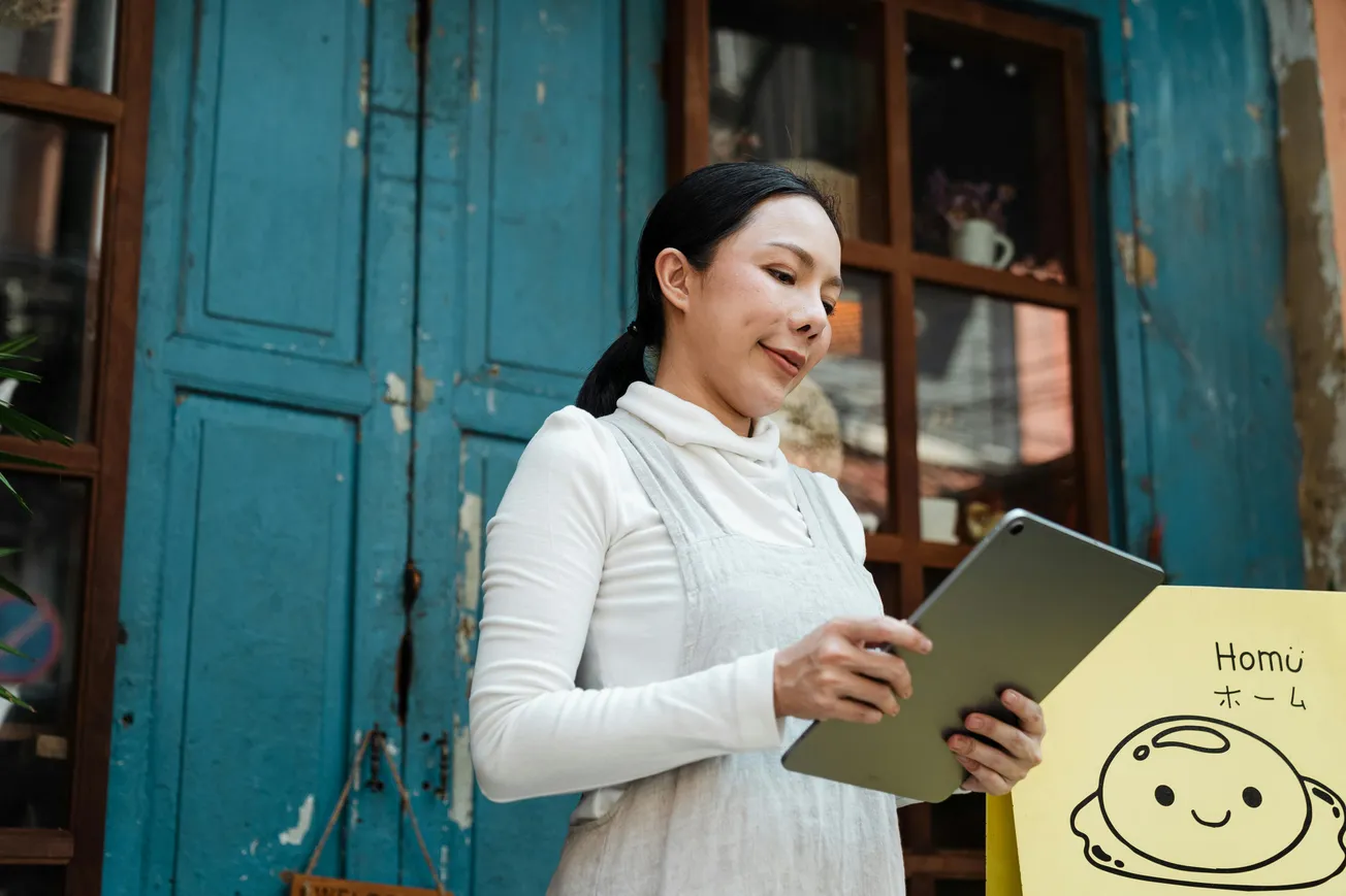 A woman stands smiling, holding a tablet in front of a weathered blue door. A cute illustrated sign with a smiling face is beside her, creating a cheerful ambiance.