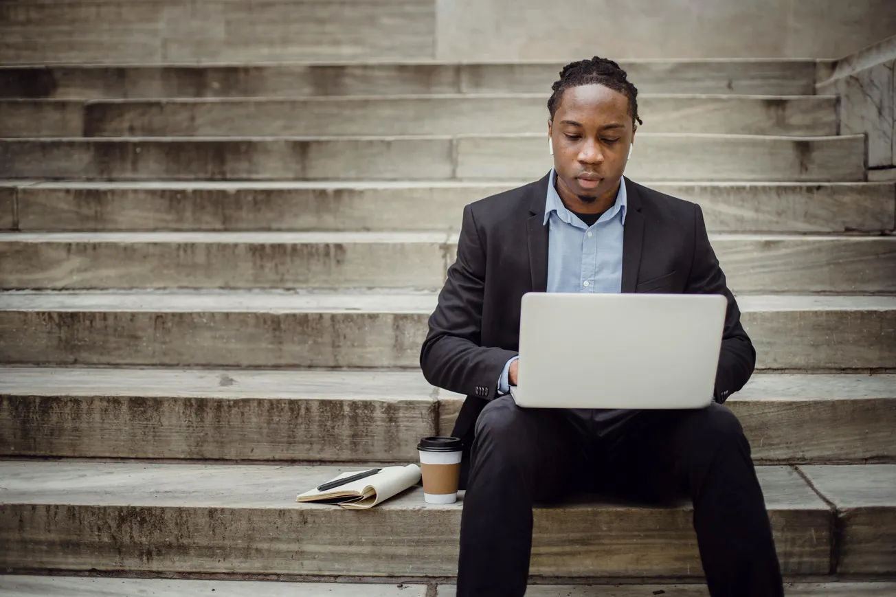 A man in a suit sits on stone steps working on a laptop, exuding focus. Beside him are a coffee cup, notebook, and pen, suggesting a professional setting.