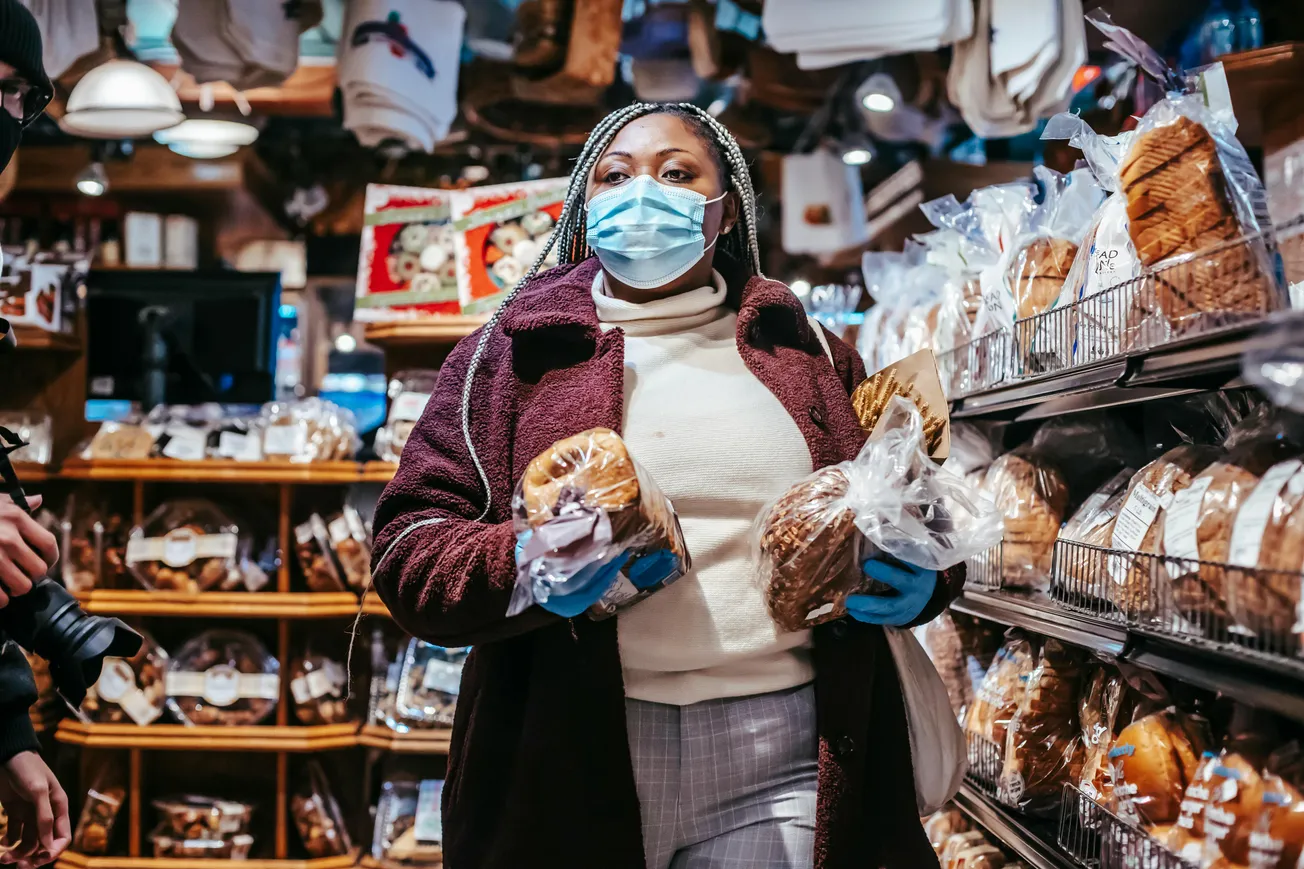 A woman wearing a face mask and gloves shops for bread in a cozy bakery. Shelves filled with packaged loaves surround her, conveying a sense of warmth.