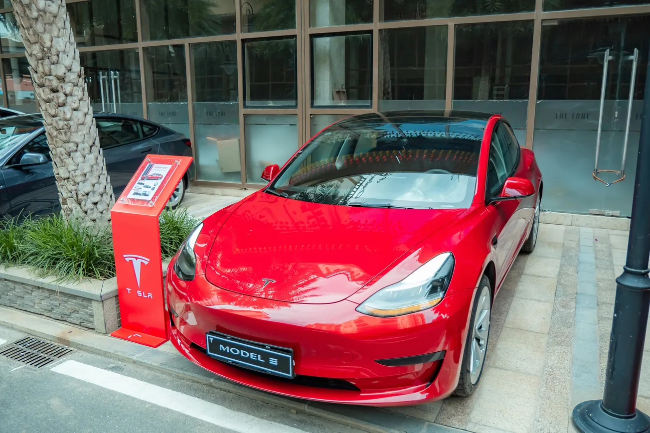 A red Tesla Model 3 parked on a city street beside a palm tree and a Tesla charging station. The background features a glass-fronted building.