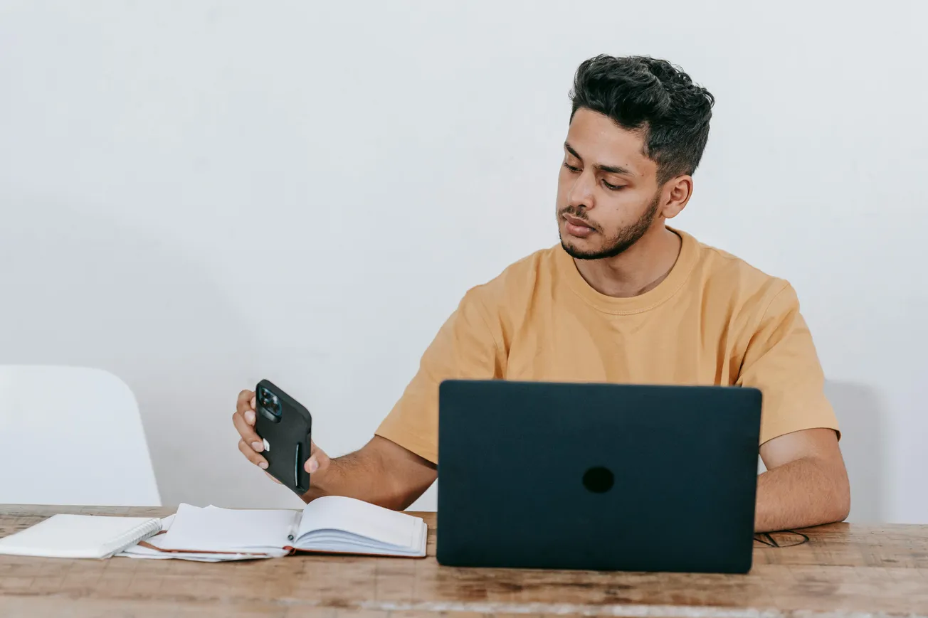 A man in a brown shirt sits at a wooden desk using a laptop, while holding a smartphone. Open notebook nearby, focusing intently, suggests productivity.