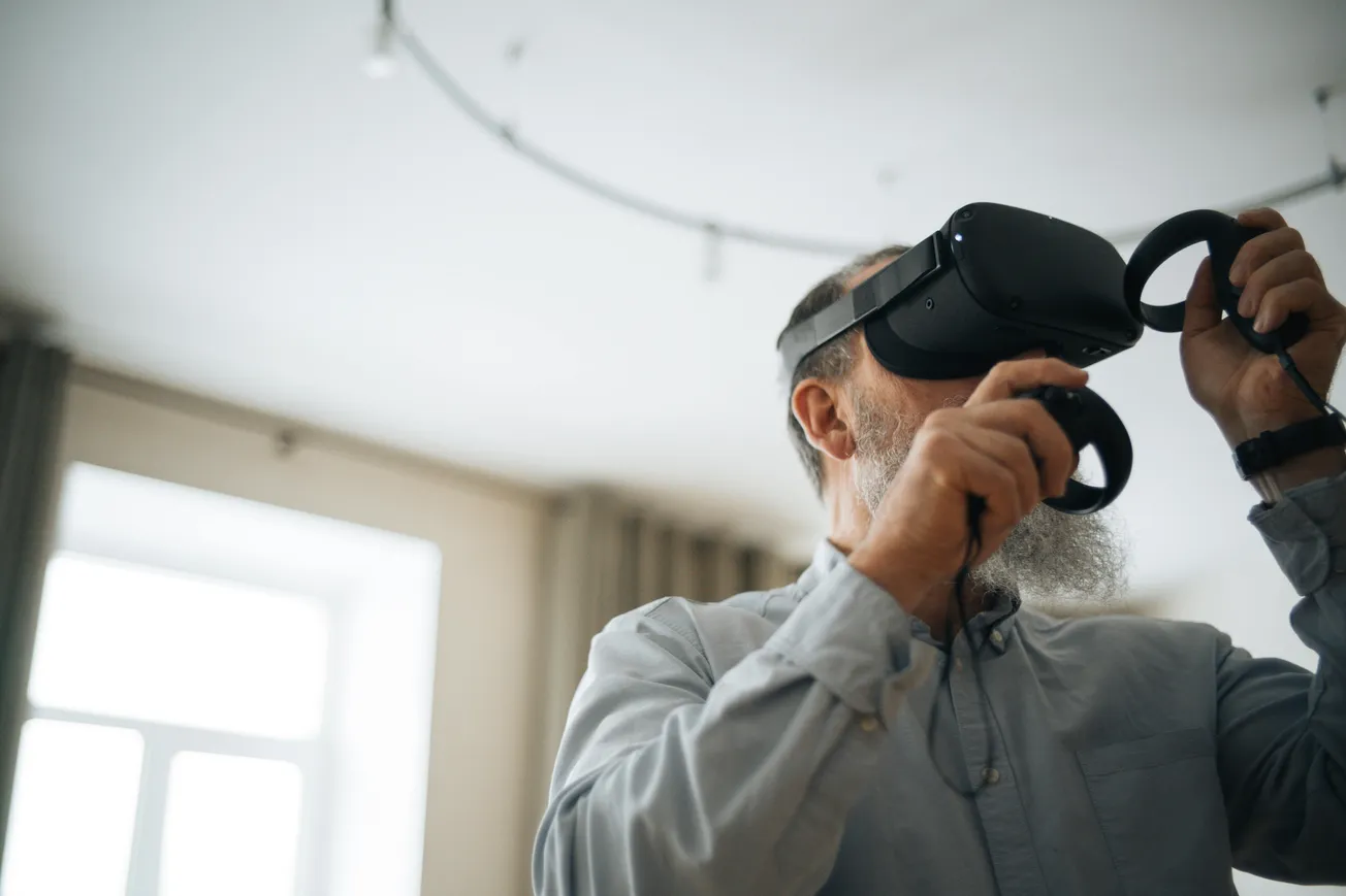 Elderly man with a gray beard uses a VR headset and controllers in a softly lit room, expressing curiosity and engagement with technology.