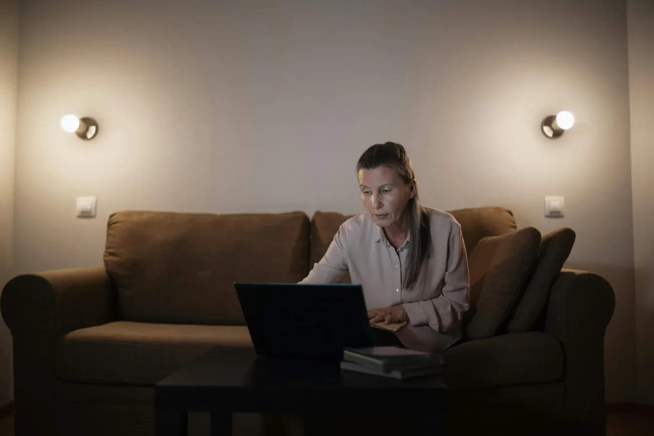 An older woman in a dimly lit room sits on a brown sofa, focused intently on her laptop. Two wall lights create a calm, serene ambiance.