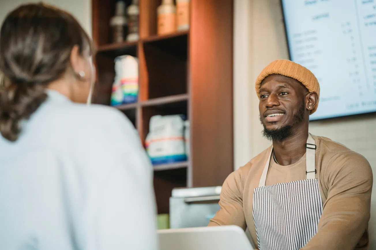 A barista wearing a mustard beanie and striped apron smiles while speaking to a customer. Shelves with colorful items and a menu screen are behind them.