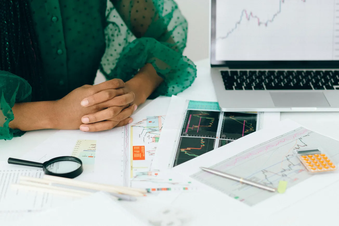 A person in a green blouse sits at a desk, hands clasped. Charts, graphs, a calculator, and a laptop display financial data, suggesting analysis.