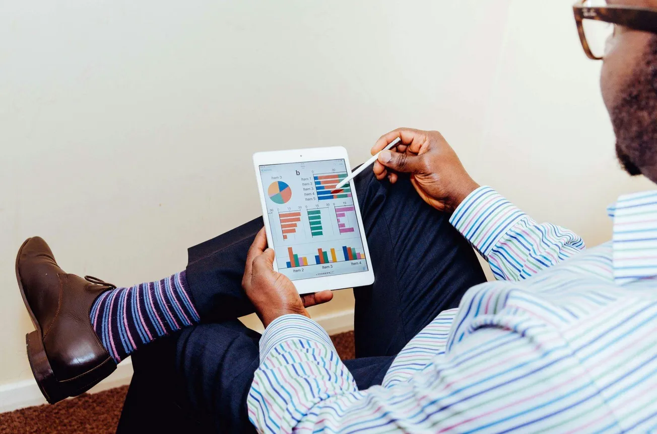 Man in striped shirt, sitting with legs crossed, viewing colorful charts on a tablet, using a stylus. Relaxed and focused atmosphere.