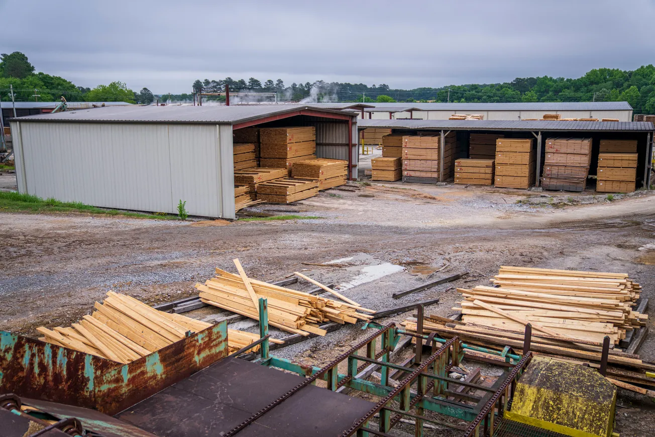 Stacks of neatly cut wooden planks are stored under metal sheds in a lumber yard. The scene is overcast, with a gravel path and green trees in the background.