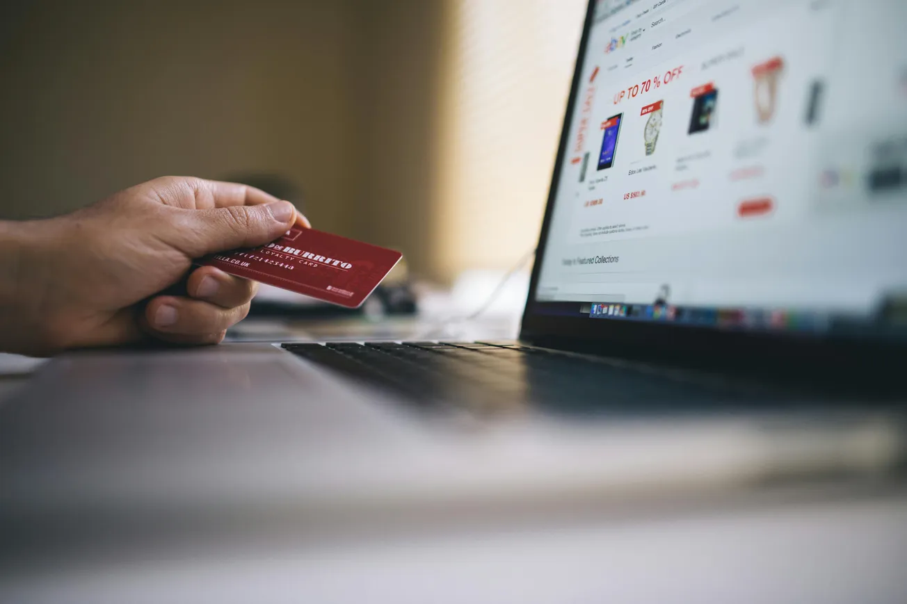 A hand holds a red credit card in front of an open laptop displaying an online shopping site. The scene suggests online shopping or e-commerce.