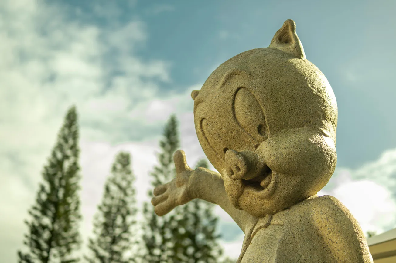 Stone statue of a cartoon character smiling and gesturing with its hand against a backdrop of tall trees and a cloudy blue sky. Playful and cheerful mood.