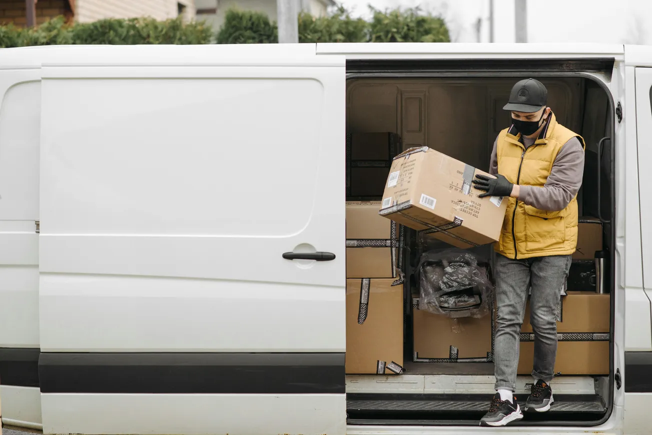 A delivery person in a yellow vest and mask unloads a large box from a white van filled with packages. The scene conveys a sense of busy efficiency.