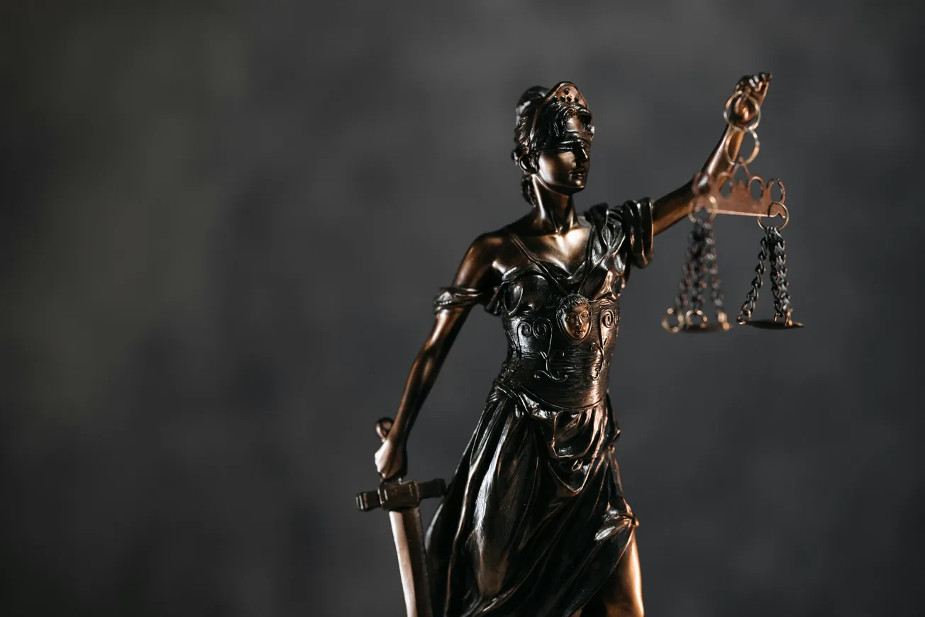 Bronze Lady Justice statue stands blindfolded, holding scales in one hand and a sword in the other against a blurred dark background, symbolizing fairness.