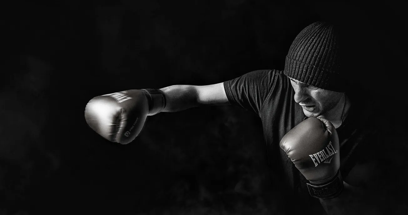 A boxer wearing a knit cap and Everlast gloves throws a punch in a dark, smoky setting. The monochrome image conveys intensity and focus.