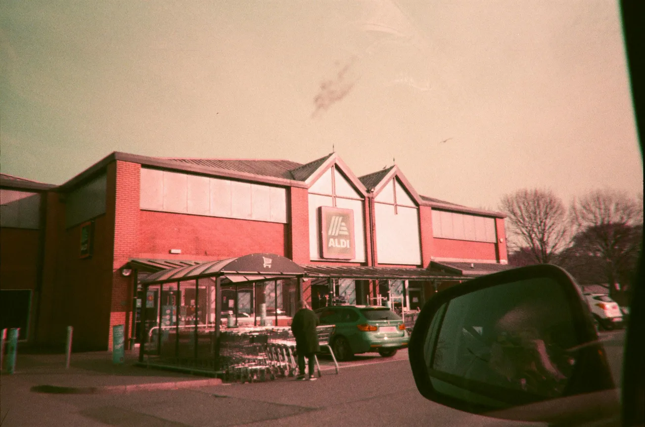 A red-brick Aldi store with a glass cart area in front. A person returns a cart beside parked cars. Trees and a car mirror frame the scene. Vintage tone.