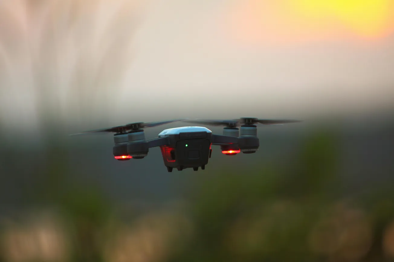 A drone with lit red lights hovers in the evening sky, silhouetted against a soft, blurred sunset. The scene is calm and focused on the flying device.