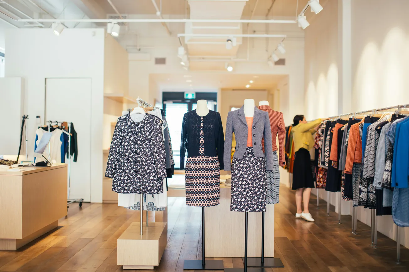 Clothing store interior with two mannequins wearing patterned dresses and cardigans. A person browses racks on the right. Bright, spacious, modern design.