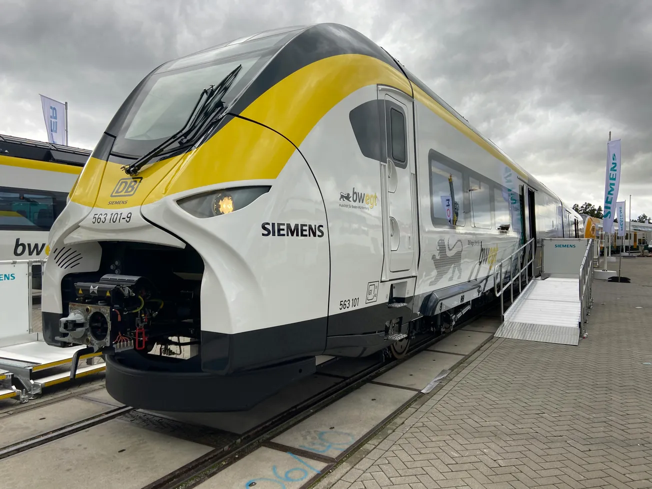 A sleek modern train with yellow, black, and white design is displayed on tracks under a cloudy sky. The train features large windows and a cockpit.