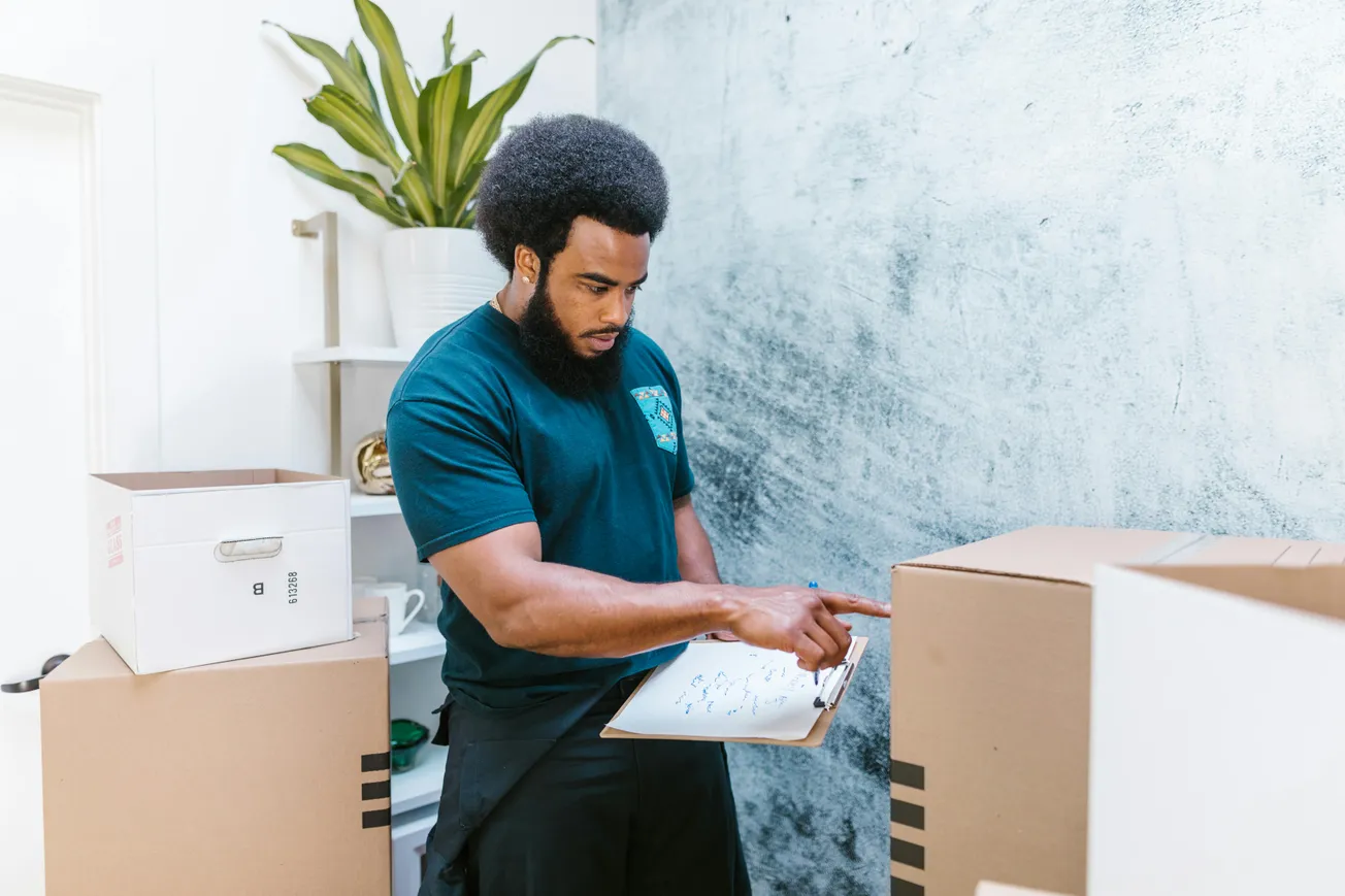 A man with an afro and beard checks a list on a clipboard while surrounded by cardboard boxes. 