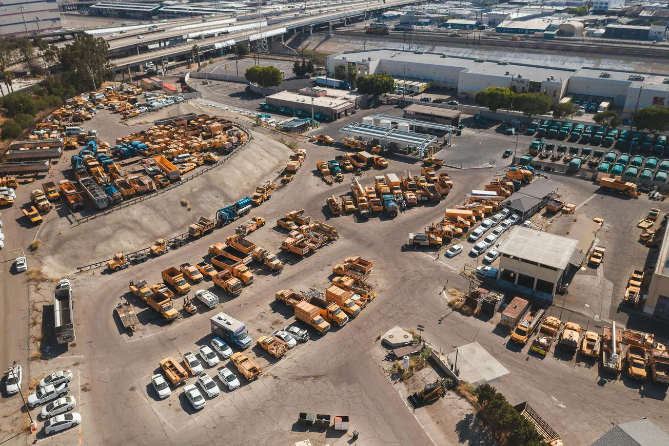 Aerial view of an industrial lot filled with organized rows of yellow trucks and equipment, white vehicles, and warehouses. The scene is orderly and busy.