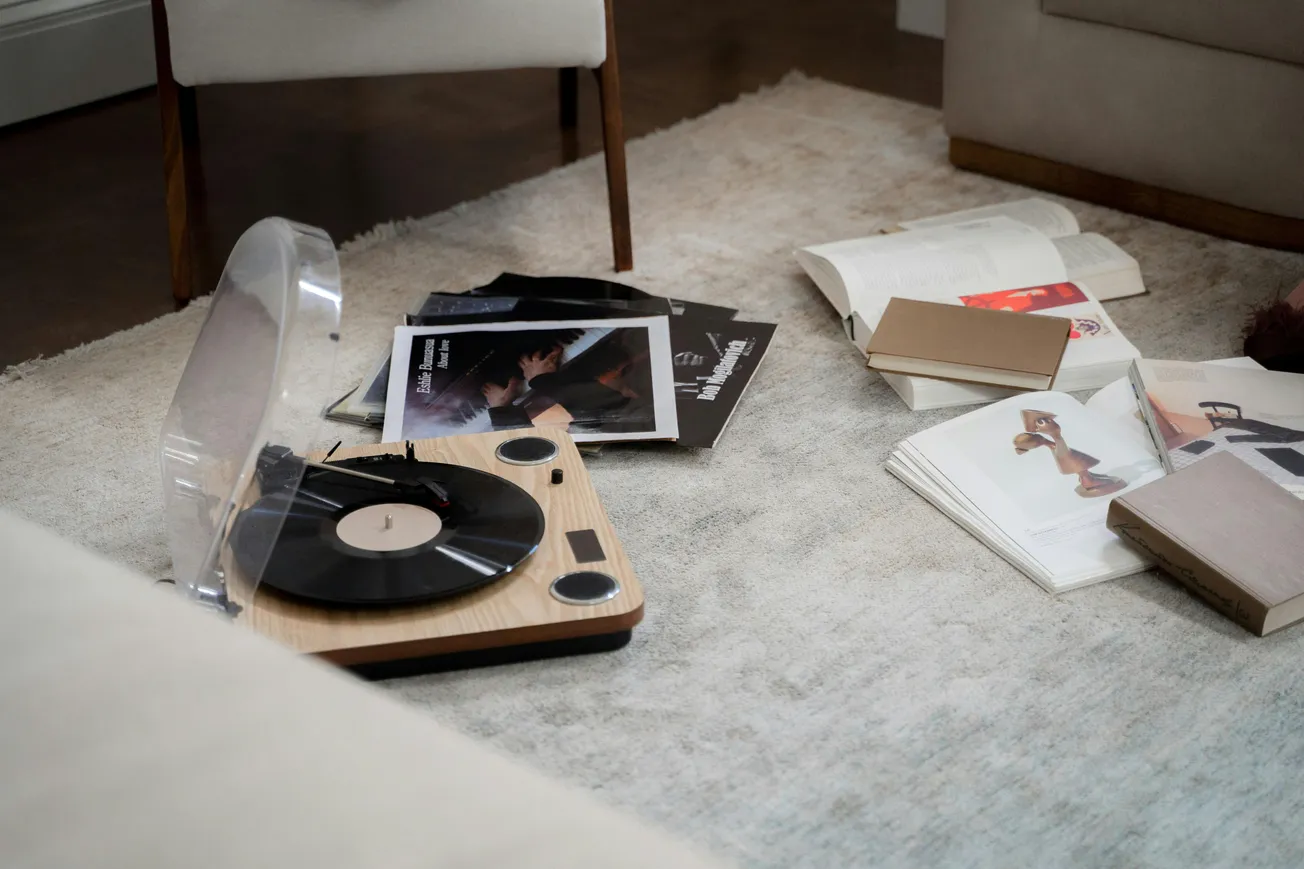 A cozy room features a vintage record player on a beige rug, surrounded by scattered vinyl covers and open magazines, conveying a relaxed, nostalgic vibe.