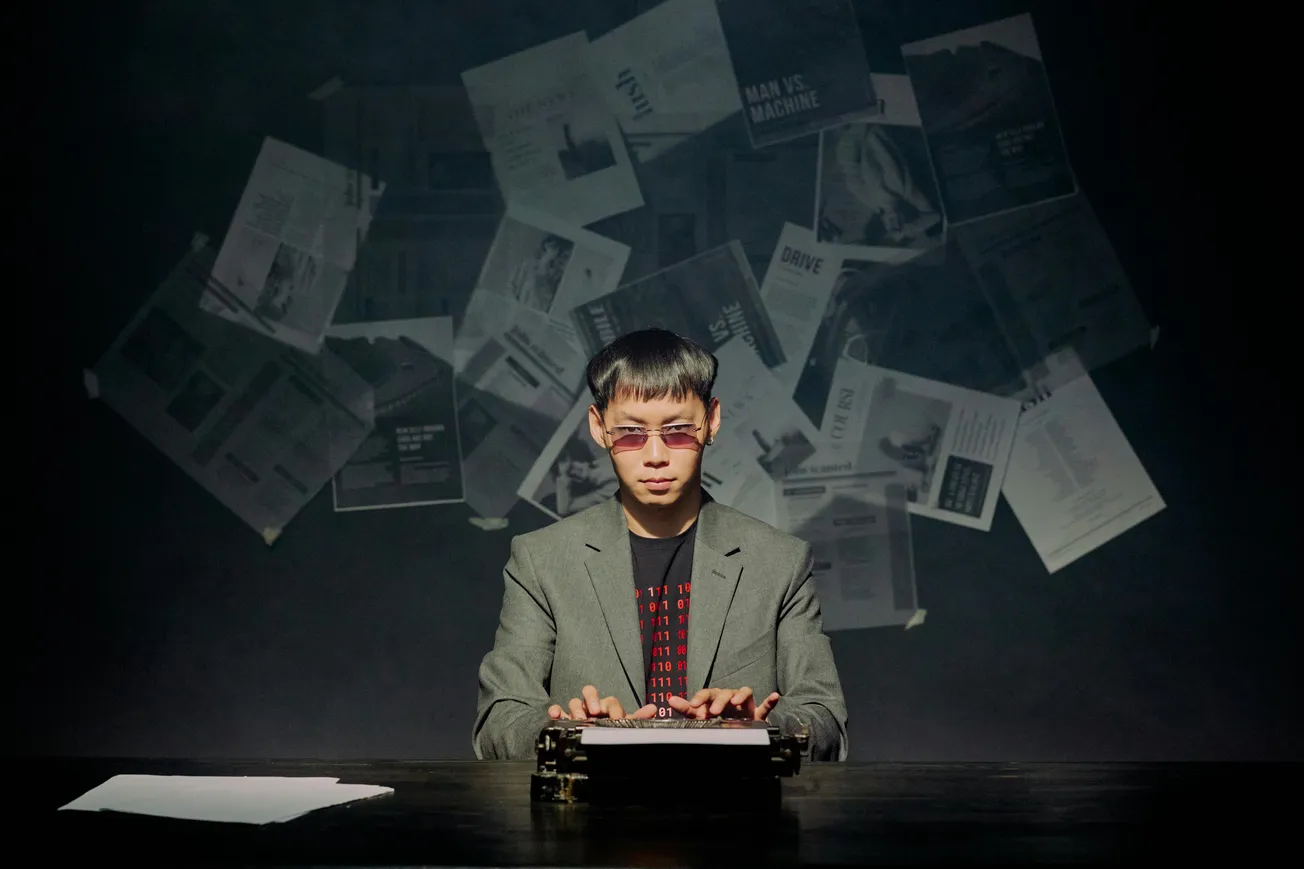 A person in a suit and sunglasses types on a typewriter against a dark background. Papers are scattered in the air, creating a mysterious, intense atmosphere.
