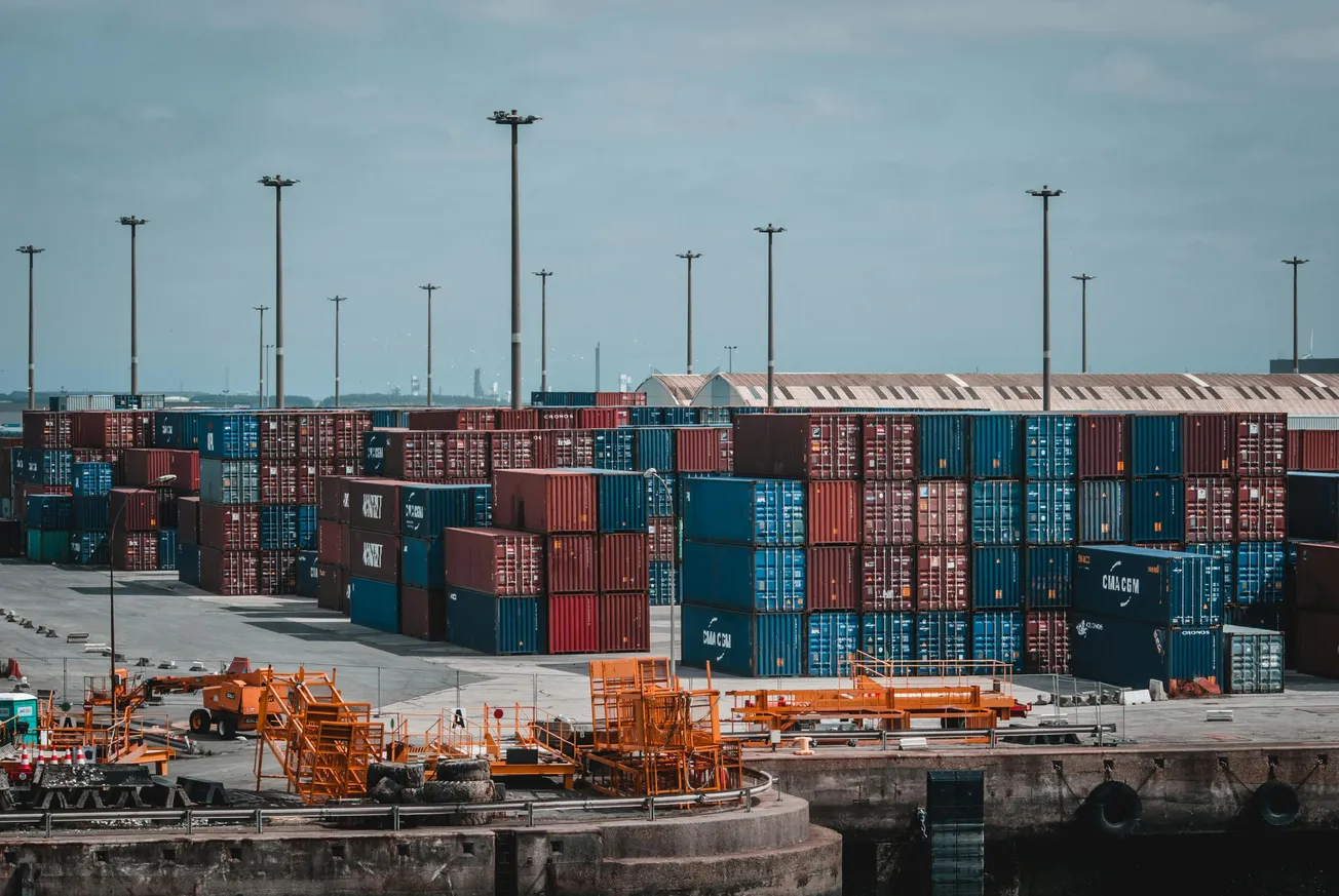 Stacks of colorful shipping containers at a busy harbor under a cloudy sky, with industrial equipment and towering light poles in the foreground.