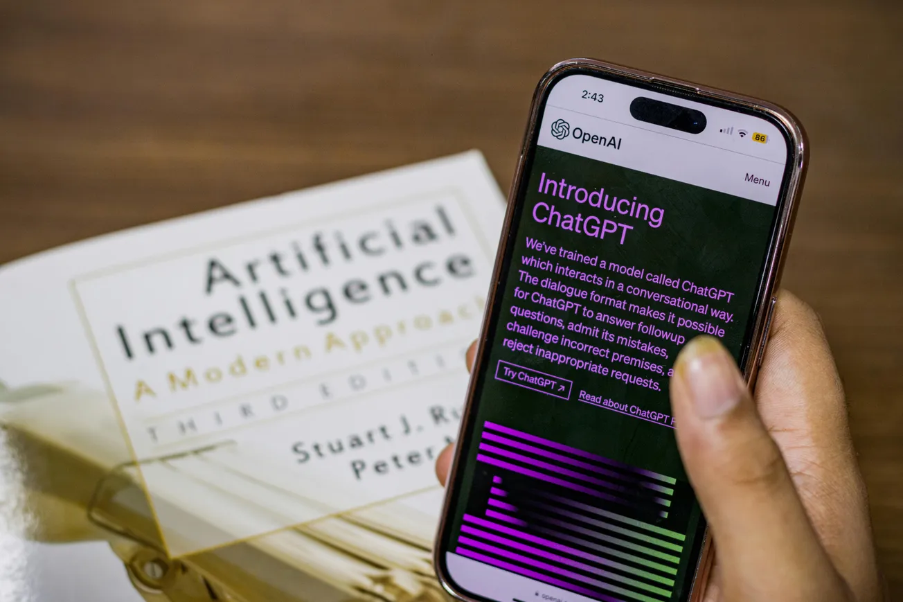 A person holds a smartphone displaying OpenAI's ChatGPT webpage. A book titled "Artificial Intelligence: A Modern Approach" lies on a wooden table.