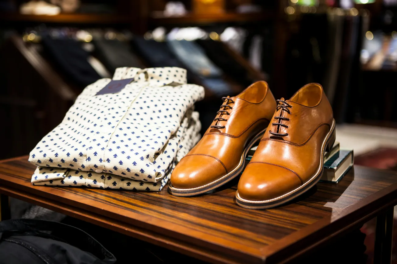 Polished brown leather dress shoes and a folded patterned white shirt with dark motifs are displayed on a wooden table, evoking a classic, elegant tone.