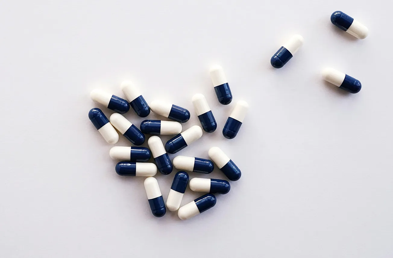 A collection of blue and white capsules scattered on a white surface, with most grouped together forming a loose heart shape, conveying a clinical tone.
