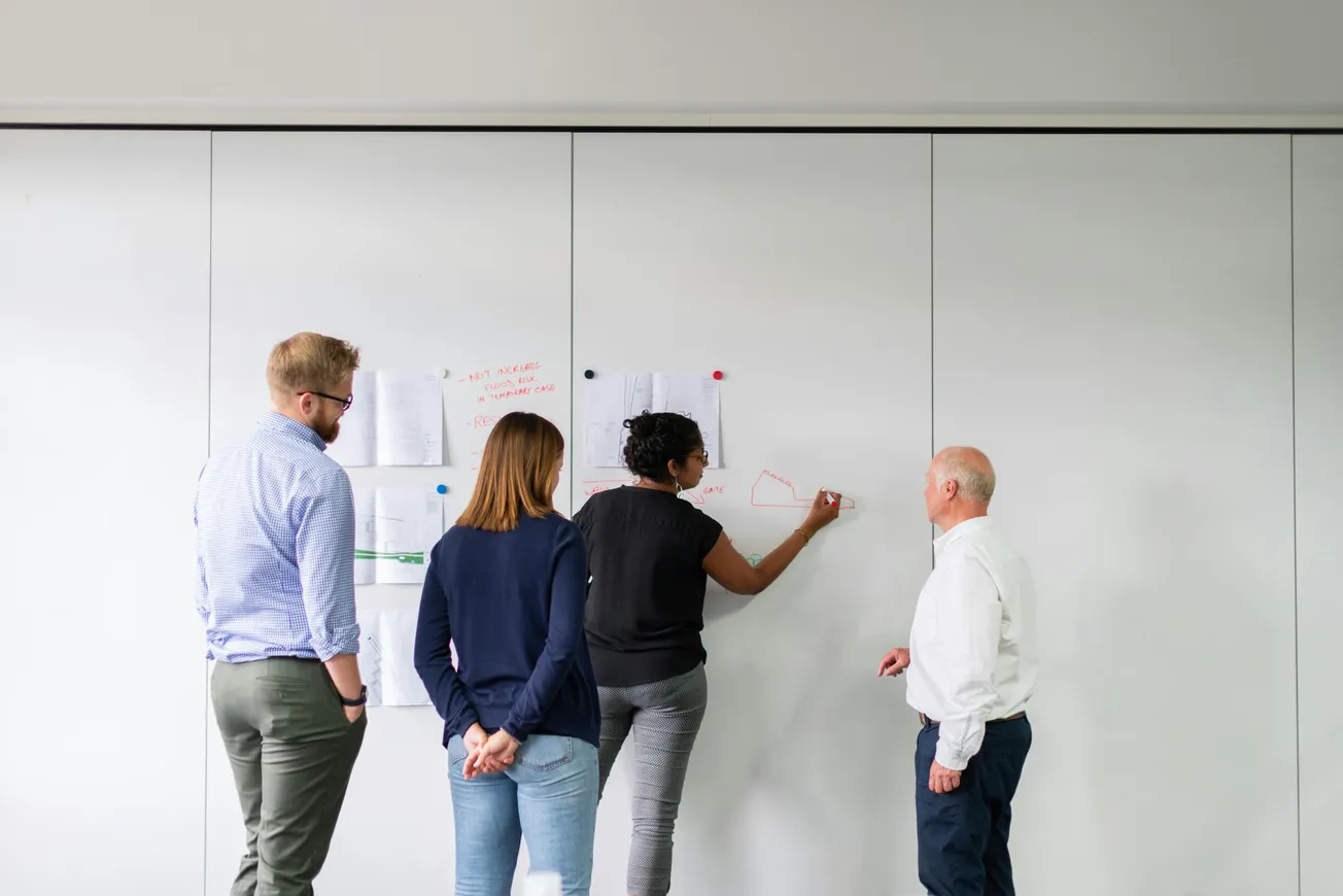 Four people collaborate in front of a whiteboard, with one writing on it. Papers with diagrams are pinned. The atmosphere is professional and focused.