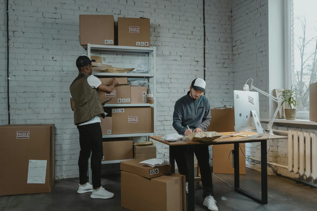 Two people in a room with a white brick wall, organizing boxes labeled "Fragile." One person stands by shelves, the other writes at a desk. The mood is focused and busy.