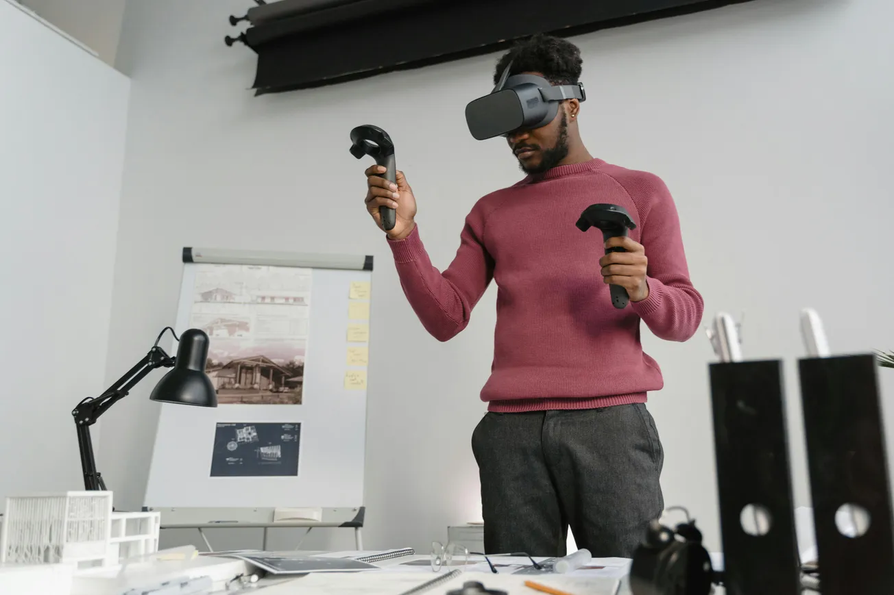 A man wearing a VR headset and pink sweater uses controllers in an office. A flipchart with architectural sketches and desk items are visible, suggesting focus.