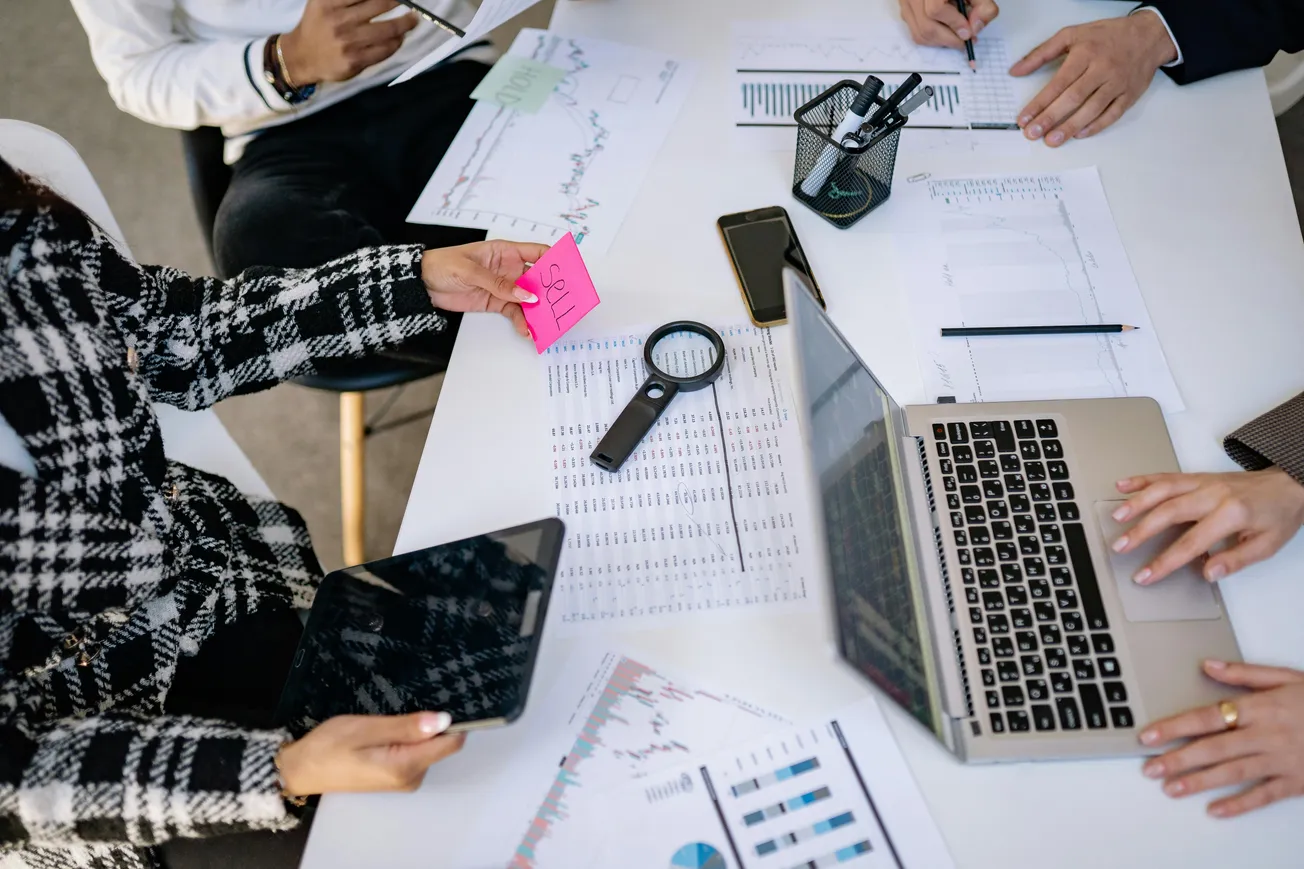 Overhead view of a collaborative office meeting. Four people around a table with charts, a laptop, magnifying glass, and notes, creating a focused, analytical atmosphere.