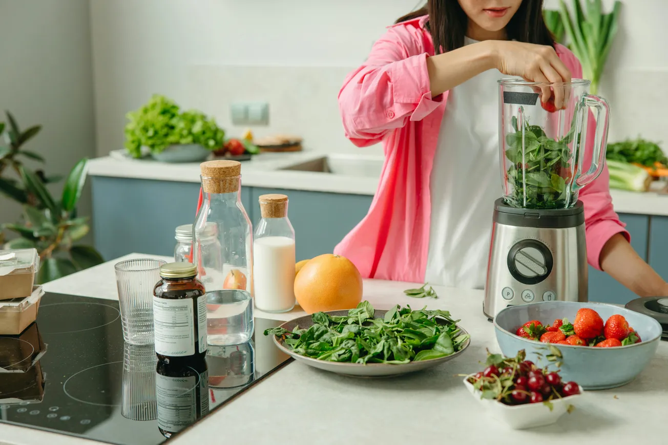 Woman in a pink shirt preparing a smoothie with fresh spinach in a blender. Kitchen counter filled with fruits, vegetables, and bottles.