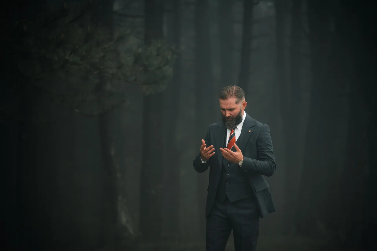 A bearded man in a dark suit with a red tie stands in a misty forest, looking contemplative with his hands raised. The mood is mysterious and introspective.