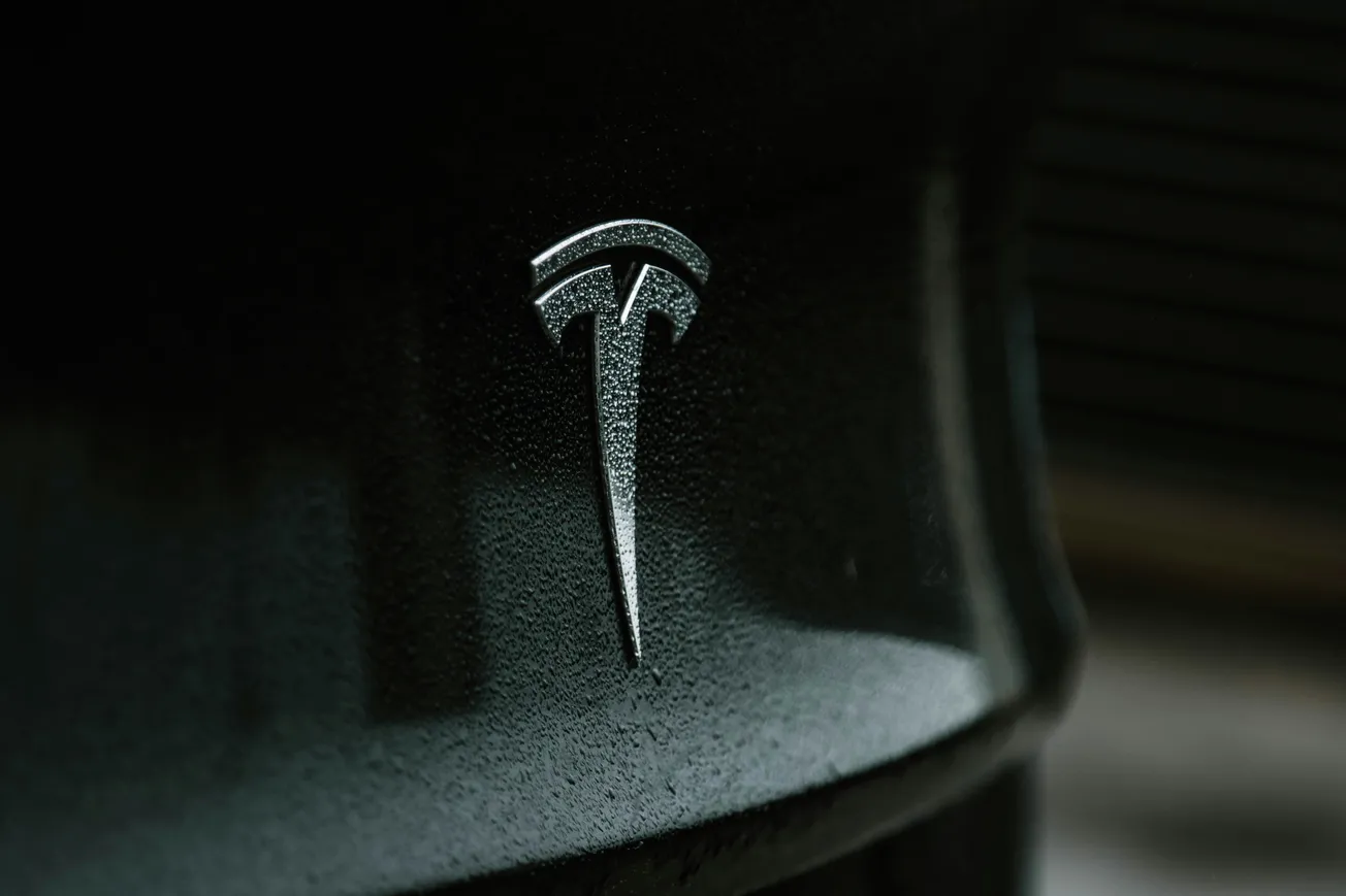 Close-up of a silver Tesla logo on a sleek, dark surface. The reflective material and soft lighting convey a modern and sophisticated feel.