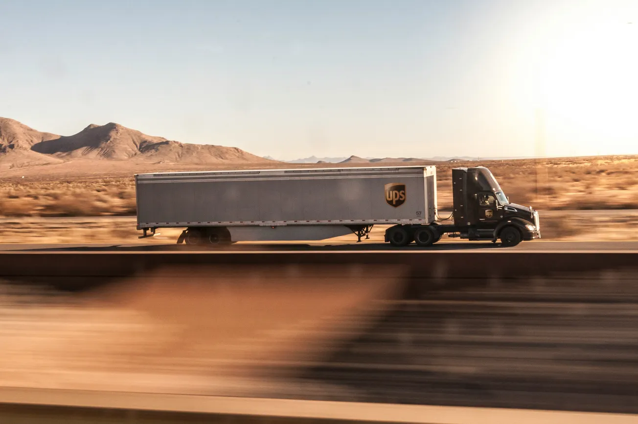 A UPS semi-truck drives along a highway through a sunlit desert landscape. Mountains are visible in the background, conveying a sense of vastness and speed.