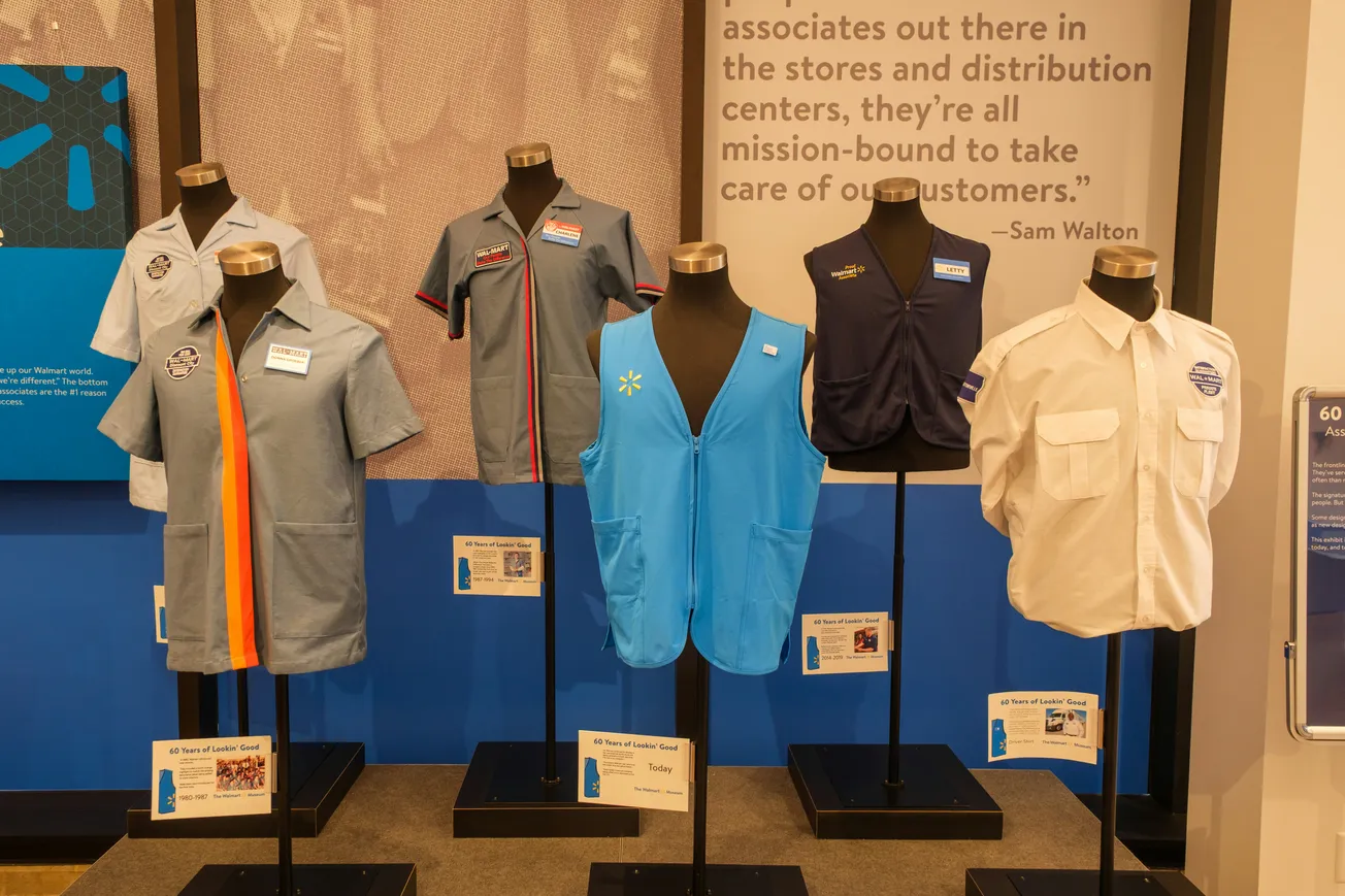 Display of six vintage Walmart uniforms on mannequins, showcasing blue, white, and gray shirts and vests against a background with a Sam Walton quote.