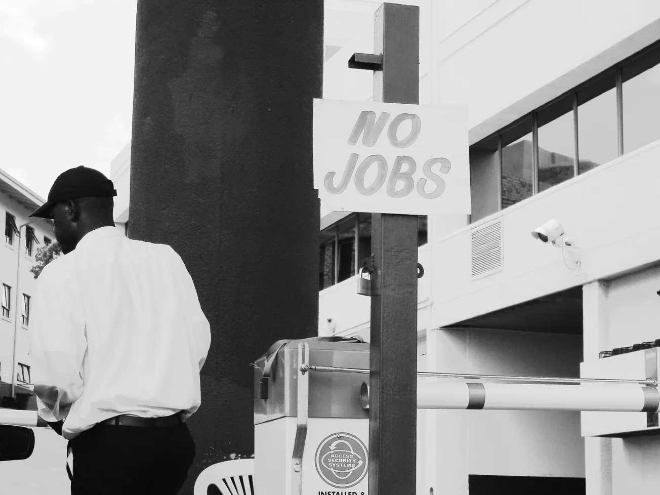 Black and white image of a man in a white shirt and cap walking near a sign reading "NO JOBS" on a post. Urban setting, conveys a somber mood.