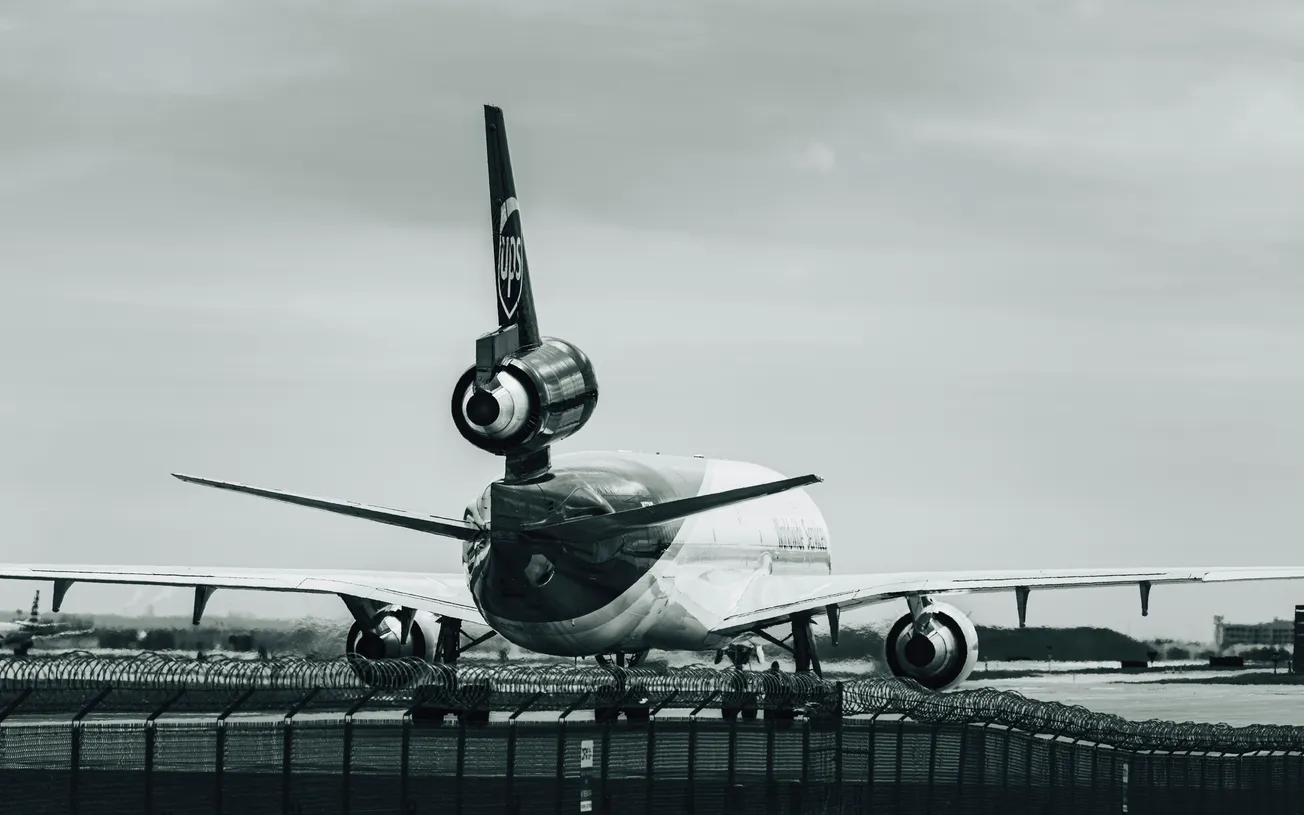 Monochrome image of a parked cargo aircraft, viewed from the rear, on an airport runway. Overcast sky above, with a distant landscape visible. Calm atmosphere.